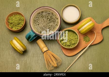 Messlöffel, grünes Matcha-Pulver und Makronen-Kekse auf Holzbrett. Milch in der Schüssel. Matcha-Tee im Becher. Grüner Hintergrund Stockfoto