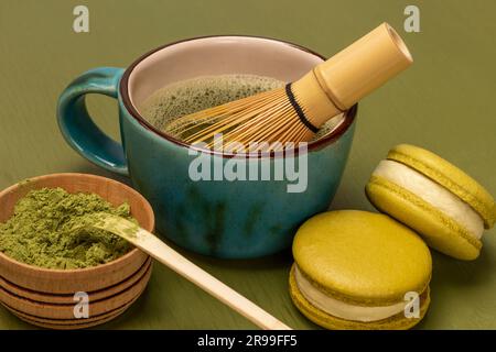 Grünes Matcha-Pulver und Messlöffel in Holzschüssel. Grüne Makronen-Kekse. Bambusquirl im Becher mit Matcha-Tee. Grüner Hintergrund Stockfoto