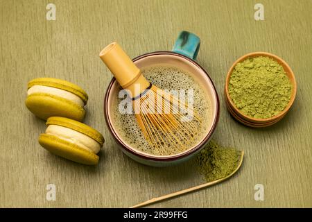 Grüne Makronen-Kekse. Bambusquirl im Becher mit Matcha-Tee. Grünes Matcha-Pulver in einer Holzschüssel. Grüner Hintergrund Stockfoto