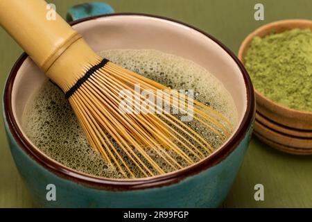 Bambusquirl im Becher mit Matcha-Tee. Grünes Matcha-Pulver in der Schüssel. Matcha-Tee in einer Holzschüssel. Grüner Hintergrund Stockfoto
