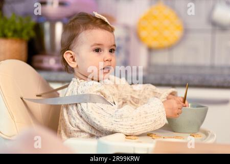 Kleines Mädchen, das in einem Hochstuhl sitzt und mit einem Löffel alleine isst Stockfoto