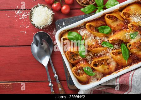 Cannelloni oder Conchiglioni. Gebackene gefüllte Nudelschalen mit Bolognese-Fleischsauce, Tomaten und Basilikum auf einem rustikalen roten Holztisch. Traditionelle italienische Fahndung Stockfoto