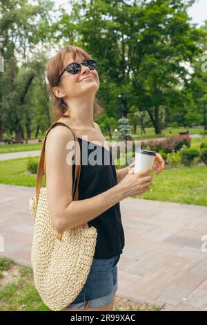 Lächelndes Mädchen mit Kaffeetasse im Park. Hübsche Frau mit Sonnenbrille, die im Park spaziert. Hintergrund für den Sommer. Sommerwochenende. Fröhlicher Freiberufler. Stockfoto