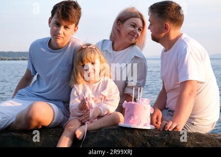 Wunderschöne Familie, verliebte Eltern, die sich gegenseitig anschauen, feiern den Geburtstag des 3-jährigen Mädchens an der Küste des Ozeans, Teenager, Junge und Kind, Baby sitzen auf Stein, schauen Sie in den Rahmen, Kinder und Erwachsene Stockfoto