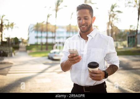 Glücklicher Geschäftsmann, der in der Pause unterwegs unterwegs mit dem Smartphone Kaffee trinkt, freier Platz Stockfoto