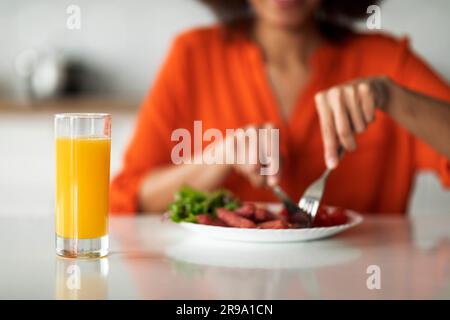 Unbekannte schwarze Frau, die Gabel und Messer benutzt, während sie in der Küche zu Mittag isst Stockfoto