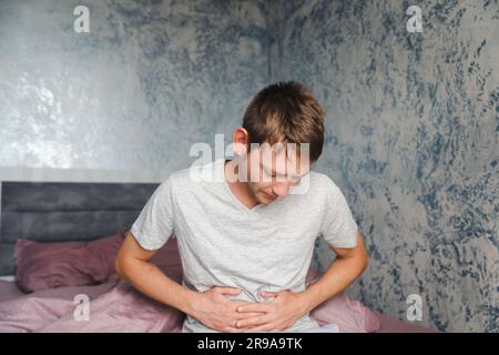 Junger weißer Mann, der unter Magenschmerzen leidet, während er zu Hause auf der Couch sitzt und seinen Bauch berührt. Männlich mit Bauchschmerzen. Das Magengeschwür Stockfoto