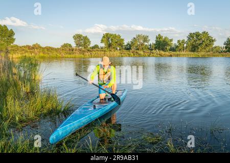 Senior männlicher Stehpaddler landet auf einem grasbedeckten Seeufer, sommerliche Landschaft in Colorado Stockfoto