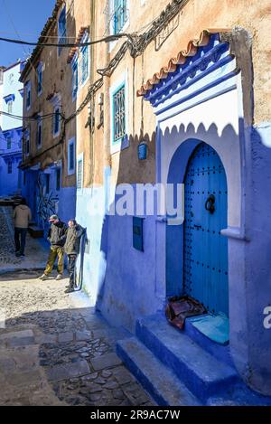 Eine dramatische, große blaue Tür im Schatten des Sonnenuntergangs mit einer typischen kleinen kopfsteingepflasterten Straße mit einheimischen Männern, die im Chefchaouen, Marokko, herumhängen Stockfoto