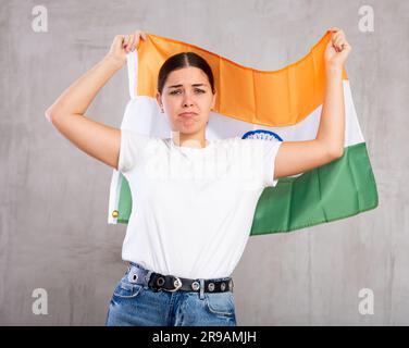 Aufgewühlte junge Frau, die die Flagge Indiens auf grauem Hintergrund hält, trauriger Ausdruck Stockfoto