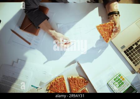 Nachhaltiger Arbeitsplatz. Draufsicht auf weibliche Mitarbeiterin im grünen Büro mit Pizza und Laptop. Stockfoto
