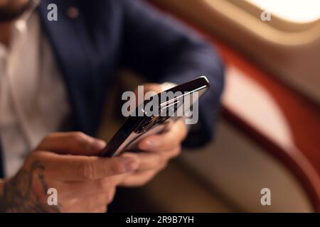 Nicht wiedererkennbarer eleganter junger asiatischer CEO Geschäftsmann mit Brille und Anzug Mann sitzt in einem erstklassigen Privatjet, während er mit einem Smartphone arbeitet Stockfoto