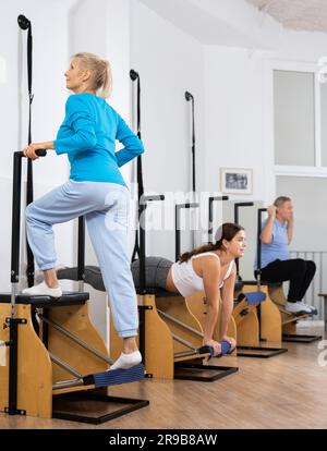 Eine entspannte Frau im Alter, die Sportbekleidung trägt und Stehübungen auf einem Pilates-Sesselautomaten im Rehabilitationszentrum macht Stockfoto