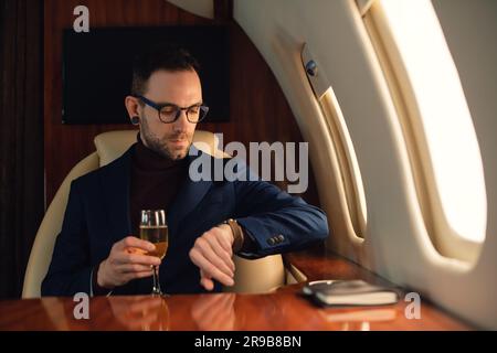 Eleganter erwachsener Geschäftsmann in einer Brille in einer blauen Jacke, während er die Zeit auf einer Armbanduhr überprüft, bevor er in einem Privatflugzeug abhebt Stockfoto