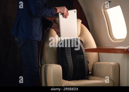 Ein eleganter Geschäftsmann aus der Nähe zieht einen modernen Laptop heraus und stellt ihn auf den Tisch, um von dort aus in einem erstklassigen Privatflugzeug zu arbeiten Stockfoto