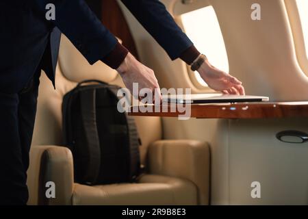 Ein unerkannter Geschäftsmann aus der Nähe zieht einen modernen Windows-Laptop heraus und stellt ihn auf den Tisch, um in einem erstklassigen Privatflugzeug zu arbeiten Stockfoto