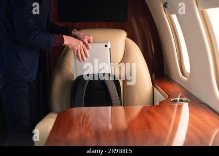 Ein eleganter Geschäftsmann aus der Nähe zieht einen modernen Laptop heraus und stellt ihn auf den Tisch, um von dort aus in einem erstklassigen Privatflugzeug zu arbeiten Stockfoto