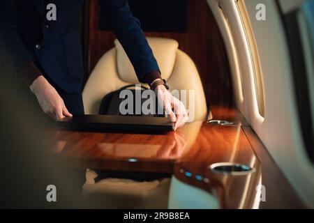 Ein unerkannter Geschäftsmann aus der Nähe zieht einen modernen Windows-Laptop heraus und stellt ihn auf den Tisch, um in einem erstklassigen Privatflugzeug zu arbeiten Stockfoto