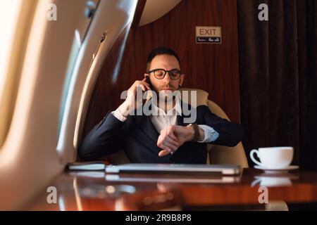 Eleganter Geschäftsmann mit Brille, der auf einem Mobiltelefon spricht, während er die Zeit vor dem Abflug auf einer Armbanduhr in einem Privatflugzeug überprüft Stockfoto