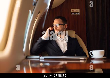 Eleganter junger Geschäftsmann mit Brille, der vor dem Start auf einem Smartphone spricht, in einem privaten Flugzeug, Reise- und Flugkonzept Stockfoto