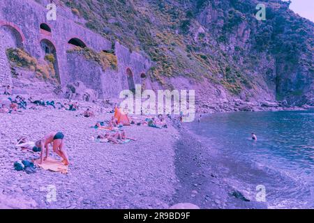 Riomaggiore Italien 26 2011. April; Steinstrand mit Sonnenanbetern unterhalb der Steinmauer am Meer in blauem Farbeffekt. Stockfoto