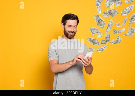 Online-Zahlung. Ein Mann kauft etwas mit einem Handy auf gelbem Hintergrund. Aus dem Gerät fliegende Dollarscheine, die den Prozess der Geldtransaktionen demonstrieren Stockfoto