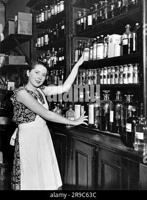 New York, New York: 1929 Marie Bard, 22, die das Modell für das 1930-Poster des Roten Kreuzes und die US-Briefmarke ist, illustriert vom Künstler Lawrence Wilbur. Sie raucht nicht, bleibt nicht lange auf und kocht und putzt für ihre Mutter, einen Apotheker und ihren Vater, einen Kaffee- und Teeimporteur, in Astoria, Queens. Hier hilft sie ihrer Mutter in der Apotheke. Stockfoto