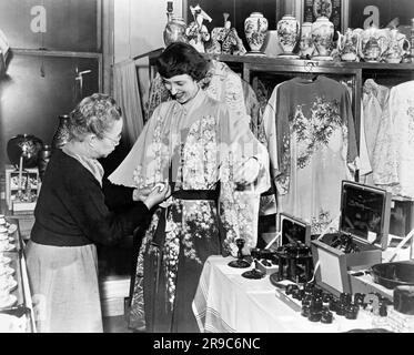 New York, New York: ca. 1955 Eine Käuferin Probiert einen Blumenmantel in einem asiatischen Geschenkeladen. Stockfoto