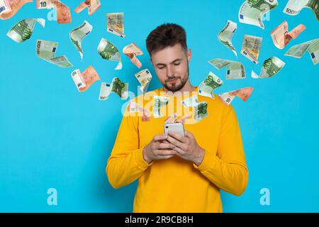 Online-Zahlung. Ein Mann kauft etwas mit einem Handy auf hellblauem Hintergrund. Aus dem Gerät fliegende Euro-Banknoten, die den Prozess des Geldes t demonstrieren Stockfoto