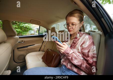Eine seriöse junge Frau, die auf dem Rücksitz eines Taxifahrzeugs sitzt und Freunden SMS schreibt Stockfoto