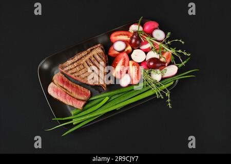 Von oben gegrillte Steaks mit drei Spitzen und Lenden, mittelschwer, mit frischem Gemüse auf schwarzem Hintergrund Stockfoto
