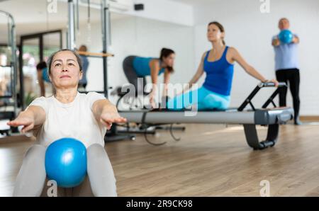 Konzentrierte ältere Frau, die während des Gruppensturses im Fitnessstudio Sit-ups mit Bändlerball macht, um die bauchmuskeln zu stärken Stockfoto