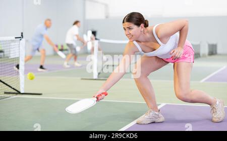 Fokussierte, emotionale, junge, weibliche Pickleball-Spielerin, die sich auf einen Schlag vorbereitet, um auf dem Hallenplatz wieder Ball zu spielen. Konzept von Wettkampfgefühlen. Stockfoto