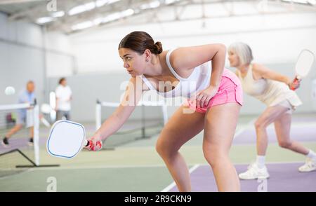 Fokussierte, emotionale, junge, weibliche Pickleball-Spielerin, die sich auf einen Schlag vorbereitet, um auf dem Hallenplatz wieder Ball zu spielen. Konzept von Wettkampfgefühlen. Stockfoto