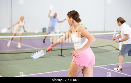 Rückansicht eines jungen Mädchens, das auf dem Hallenplatz auf einem unscharfen Hintergrund der Gegner ein Pickleball-Match spielt. Sport und aktives Lifestyle-Konzept. Stockfoto