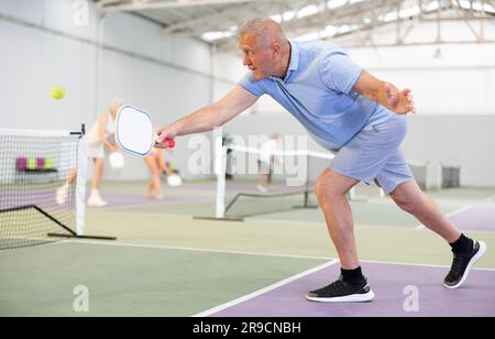 Porträt eines aktiven, emotionalen älteren Mannes, der auf dem Hallenplatz Pickleball spielt, das Paddel schwingt, um den Ball über das Netz zurückzugeben. Stockfoto