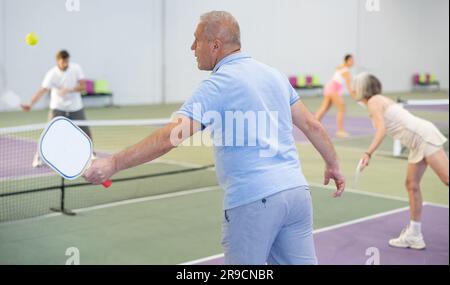 Rückansicht eines fokussierten älteren Mannes, der auf dem Hallenplatz ein Pickleball-Match mit verschwommenem Hintergrund der Gegner spielt. Sport und aktives Lifestyle-Konzept. Stockfoto