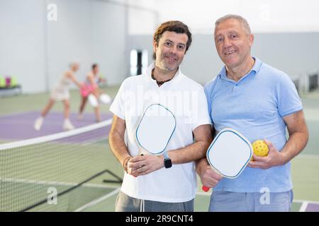 Zwei positiv lächelnde Männer in Sportbekleidung mit Schlägern und Bällen in den Händen, die nach einem freundlichen Spiel neben dem Netz auf dem Pickleball-Platz in der Halle posieren. Stockfoto