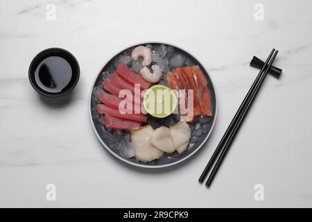 Sashimi-Set (rohe Scheiben von Thunfisch, öligem Fisch, Lachs und Garnelen) serviert mit Limette, Sojasoße und Eis auf einem weißen Marmortisch, flach liegend Stockfoto