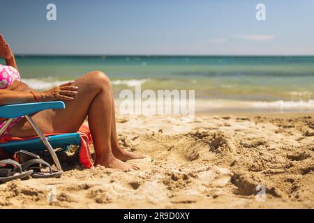 Beine einer Frau am Strand aus der Nähe, Frau sonnt sich auf einer Liege in sitzender Position am Strand Stockfoto