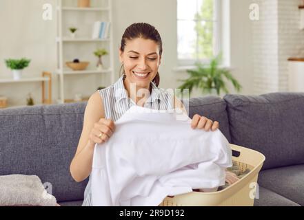 Glückliche junge Hausfrau, die ihre Wäsche überprüft, sich ein sauberes, weißes Hemd ansieht und lächelt Stockfoto