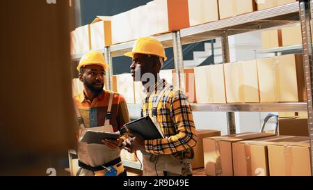 afroamerikanische Männer planen den Lagertransport mit Fracht auf Regalen, scannen Barcodes von Produkten im Lagerraum. Junge Leute, die mit Scannern und Tablets arbeiten, um den Bestand zu überprüfen. Handgeführte Aufnahme. Stockfoto