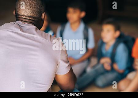 Schulbetreuer im Gespräch mit einer Gruppe von Kindern in der Grundschule. Männlicher Lehrer hält eine Motivationsrede vor seinen Schülern. Grundschulpädagoge Stockfoto