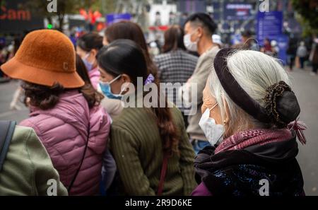 Eine ältere Frau, die traditionelle Kleidung und eine Gesichtsmaske trägt, steht neben den jüngeren Menschen in Hanoi, Vietnam. Stockfoto