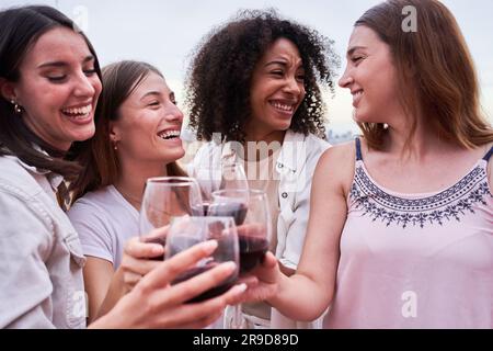 Vier gemischtrassige weibliche Freunde, die gemeinsam Spaß haben und draußen mit Rotwein anstoßen. Stockfoto