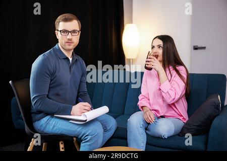 Ein junger Psychologe führt eine Psychotherapiesitzung für eine Frau mit psychischen Problemen durch. Eine Frau sitzt auf dem Sofa und hält ein Glas Wasser hinein Stockfoto
