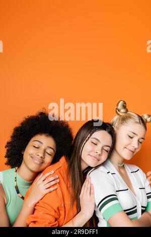 Positive und stilvolle, multiethnische Teenager-Freundinnen mit farbenfrohem Make-up, die lässige Outfits tragen und sich mit geschlossenen Augen umarmen Stockfoto