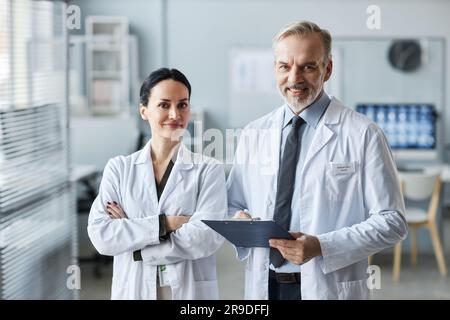 Zwei reife, erfolgreiche Hausärzte in Laborkitteln schauen in der Arztpraxis auf die Kamera, während einer von ihnen sich Notizen im Dokument macht Stockfoto