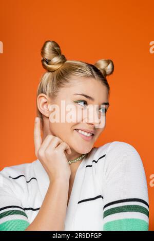 Porträt eines lächelnden und trendigen blonden Mädchens mit Frisur und auffälligem Make-up, das mit dem Finger zeigt und wegblickt, während es isoliert auf Orange steht Stockfoto