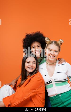 Lächelndes afroamerikanisches Teenager-Mädchen mit leuchtendem Make-up umarmt stilvolle Freundinnen in lässigen Outfits und posiert zusammen isoliert auf Orange, cool an Stockfoto